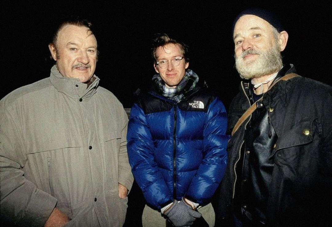 Three men stand together against a dark background. The man on the left wears a light-colored winter coat, the man in the middle wears glasses and a blue North Face puffer jacket, and the man on the right wears a dark beanie and jacket.