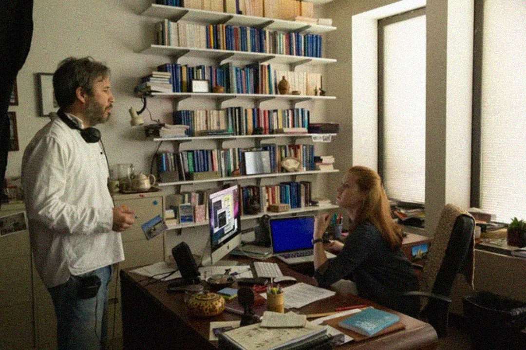 A man stands and speaks to a woman seated at a cluttered desk in an office with shelves full of books, computer monitors, papers, and large windows letting in natural light.