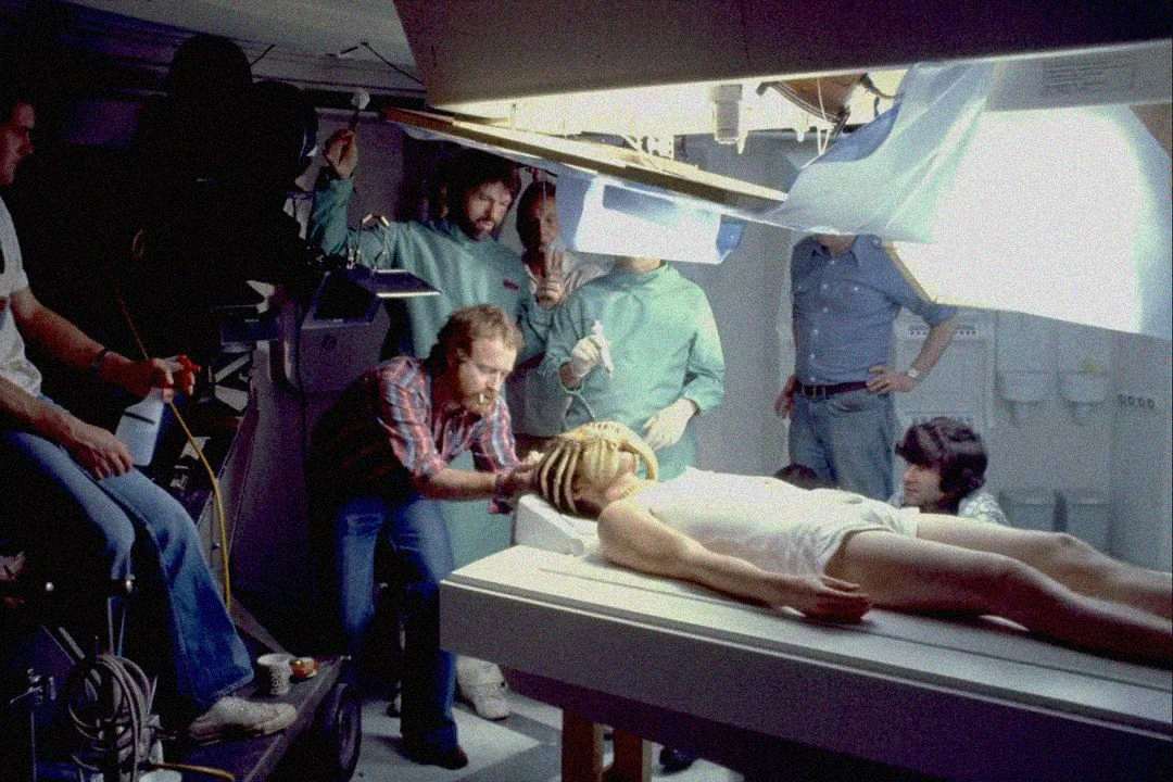 A film crew works on a set with bright lights, surrounding an actor lying on a table in a white outfit, while a crew member adjusts something near the actor’s head.