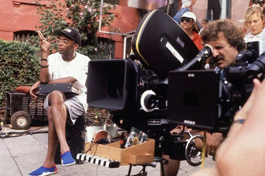 A man in casual clothing and glasses sits on a box outdoors, making a peace sign, surrounded by large film cameras and crew members during a film shoot.