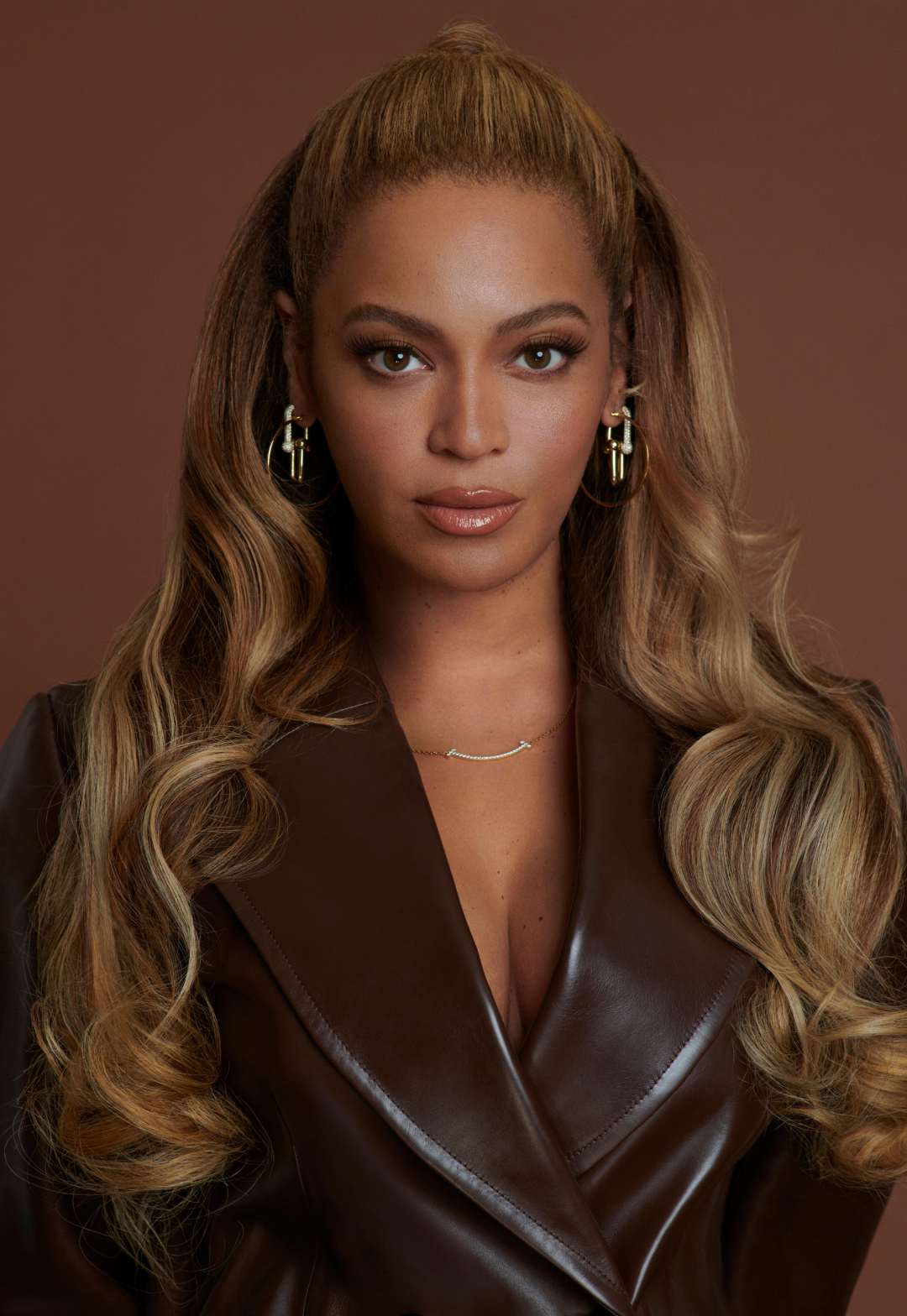 A woman with long, wavy blonde hair wears gold hoop earrings, a brown leather jacket, and a delicate necklace, posing against a solid brown background with a confident expression.
