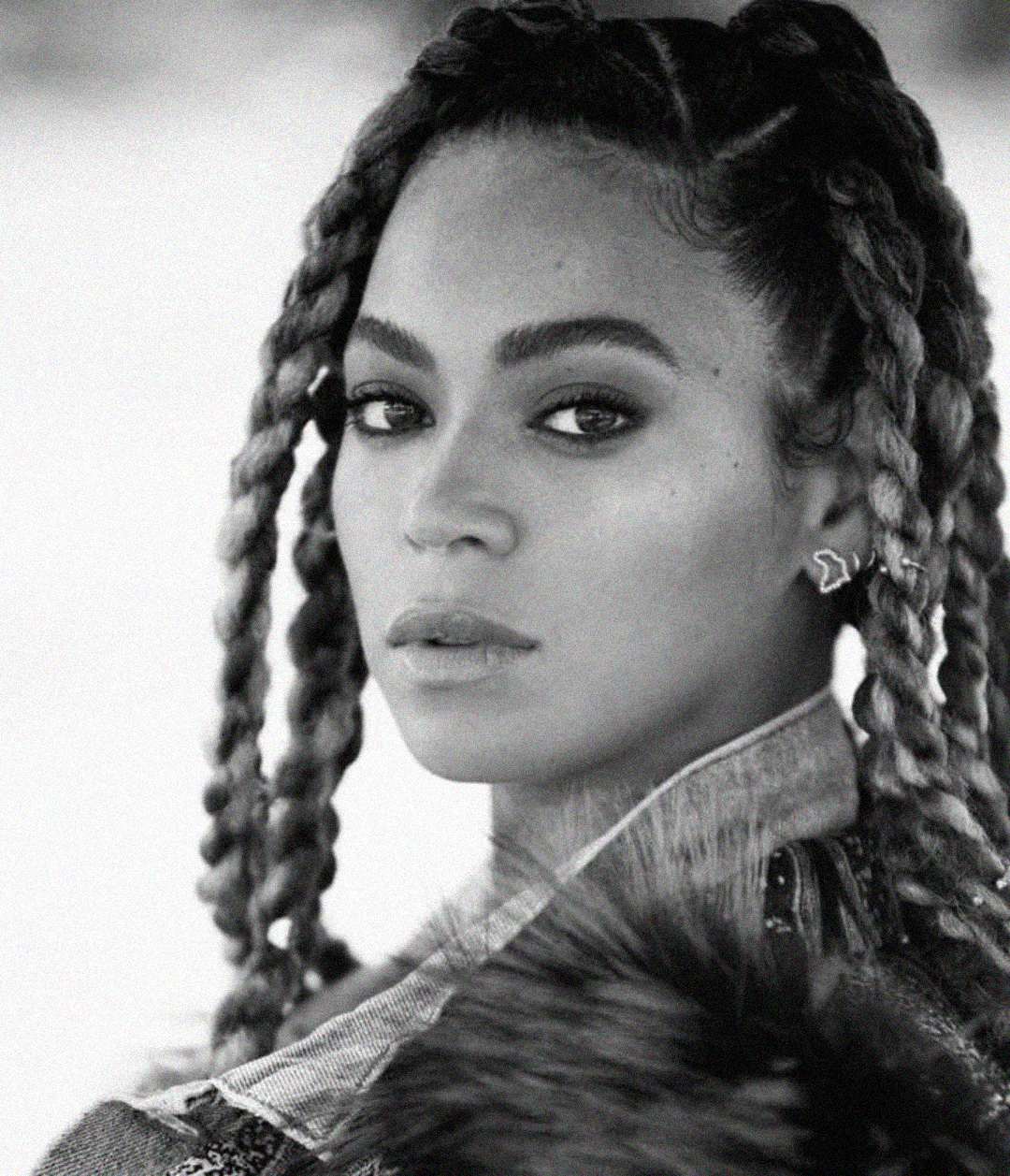 Black and white portrait of a woman with long, thick braids looking intensely at the camera. She wears a textured jacket with a fur collar and visible earrings. The background is softly blurred.