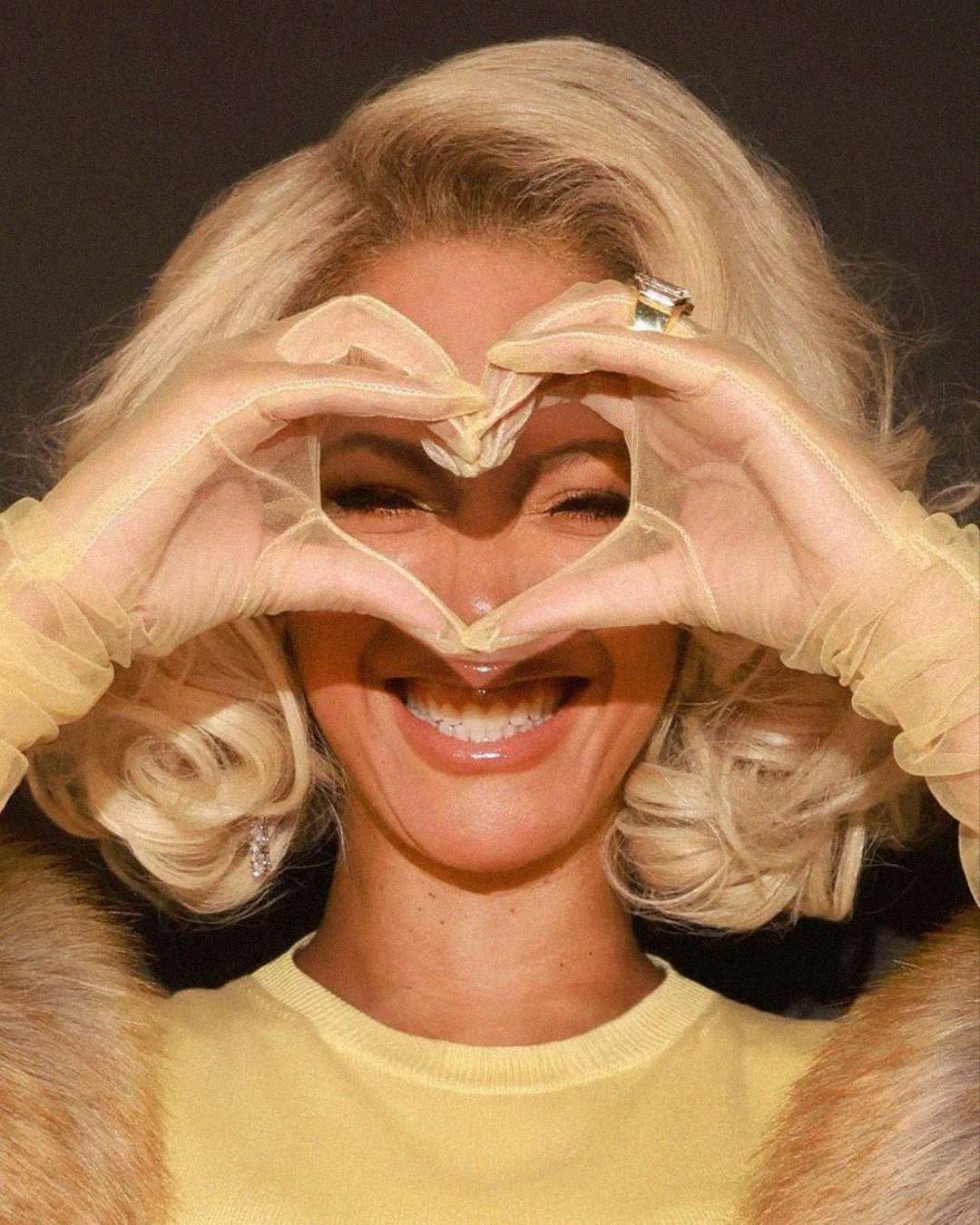 A smiling woman with blonde, curly hair forms a heart shape with her gloved hands in front of her face. She wears a yellow top, a fur stole, and large rings. The background is dark.