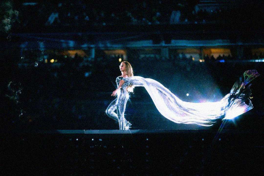 A performer in a shiny silver outfit stands onstage with a long, flowing cape trailing behind, illuminated by bright lights, while the audience watches in a darkened arena.