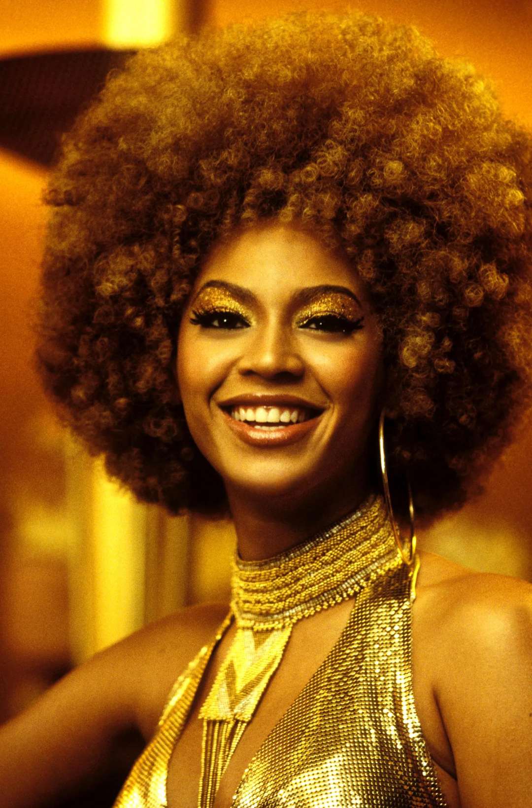A woman with a large curly afro hairstyle smiles brightly. She wears gold eye makeup, large hoop earrings, and a gold halter-neck top with matching statement jewelry. The background is warmly lit in golden tones.