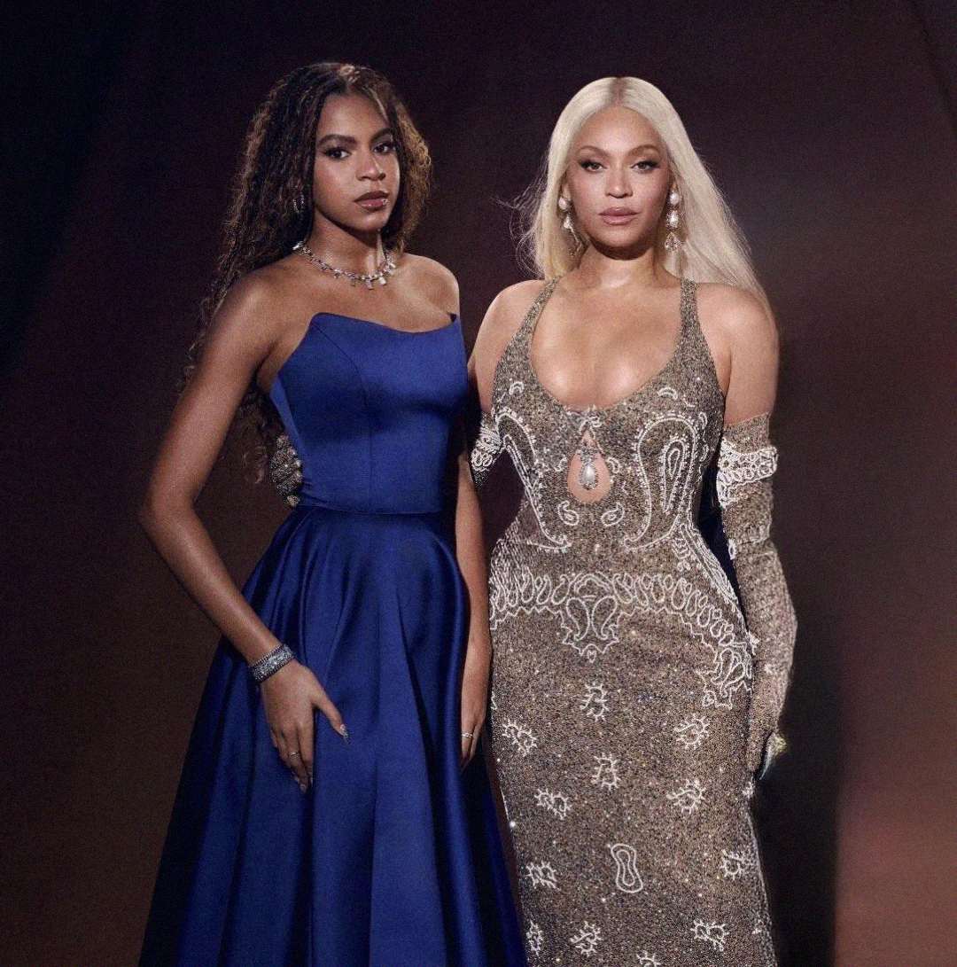 Two women stand side by side against a dark background. One wears a strapless blue gown with wavy hair, and the other wears a detailed, sparkling gown with long blond hair and matching gloves. Both look confidently at the camera.