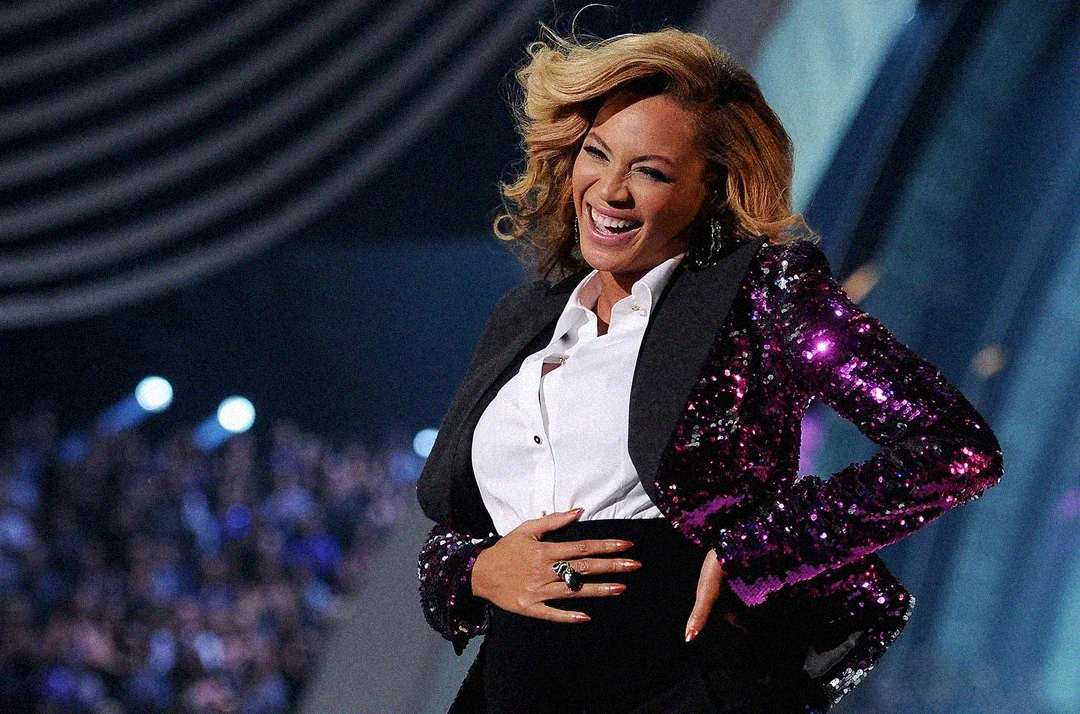 A smiling woman in a sparkling purple blazer and white shirt stands on stage, holding her stomach with both hands, with a cheering crowd and bright lights in the background.