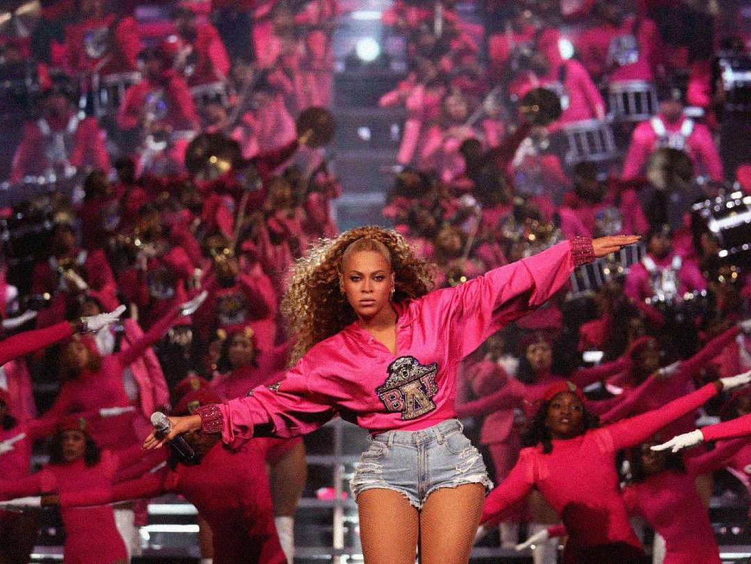 A woman in a pink top and denim shorts performs on stage, arms outstretched, with a large marching band in matching pink uniforms playing instruments in the background.