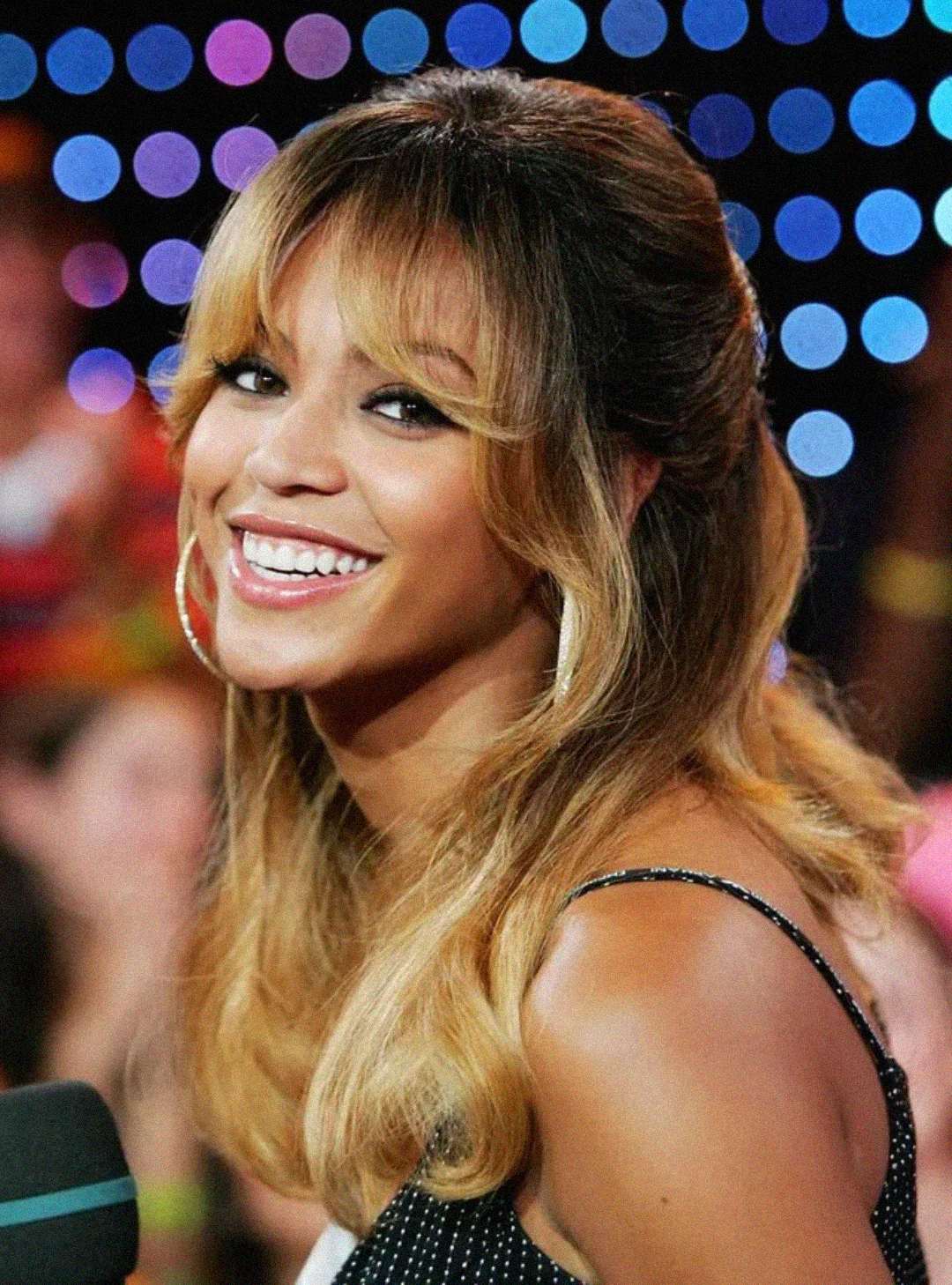 A woman with long, wavy blonde hair and bangs smiles while holding a microphone. She wears a black, sleeveless top with white polka dots. The colorful, blurred lights and people in the background suggest a lively atmosphere.