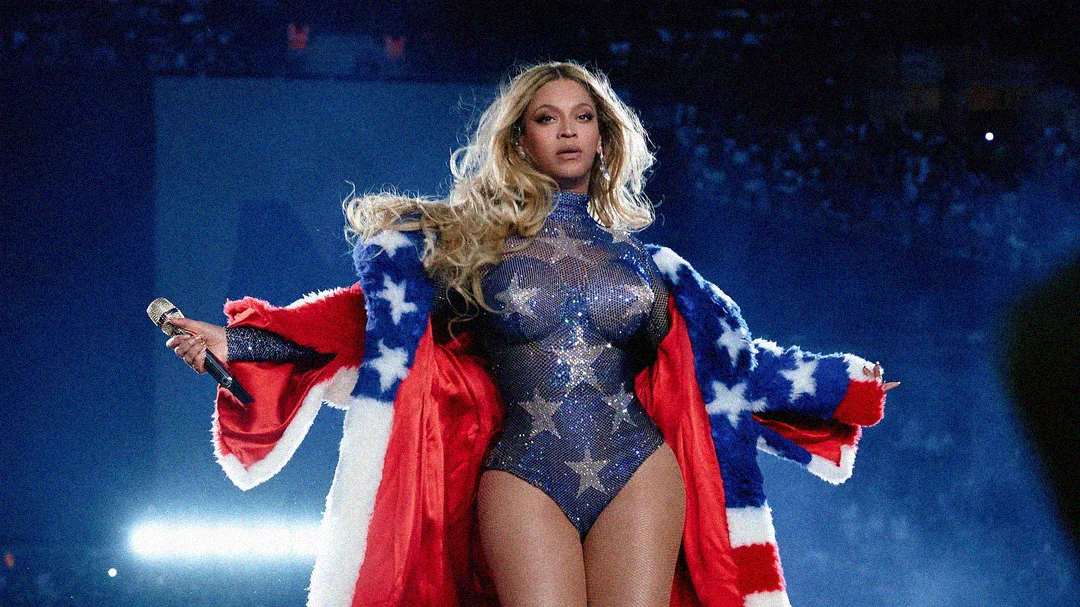 A performer wearing a sparkling bodysuit and a red, white, and blue star-spangled robe holds a microphone on stage, with dramatic lighting and a stadium audience in the background.