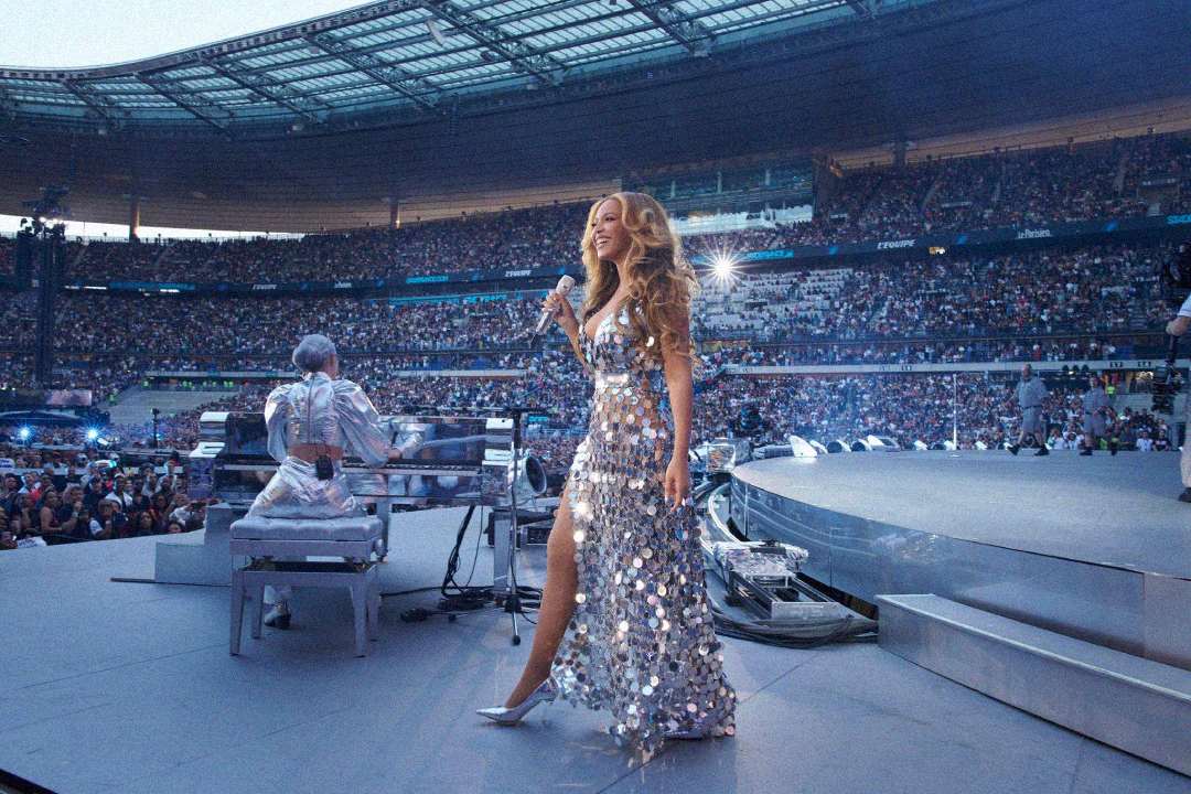 A singer in a sparkling silver gown performs on stage at a large stadium filled with a crowd, while a musician plays piano nearby under bright lights.