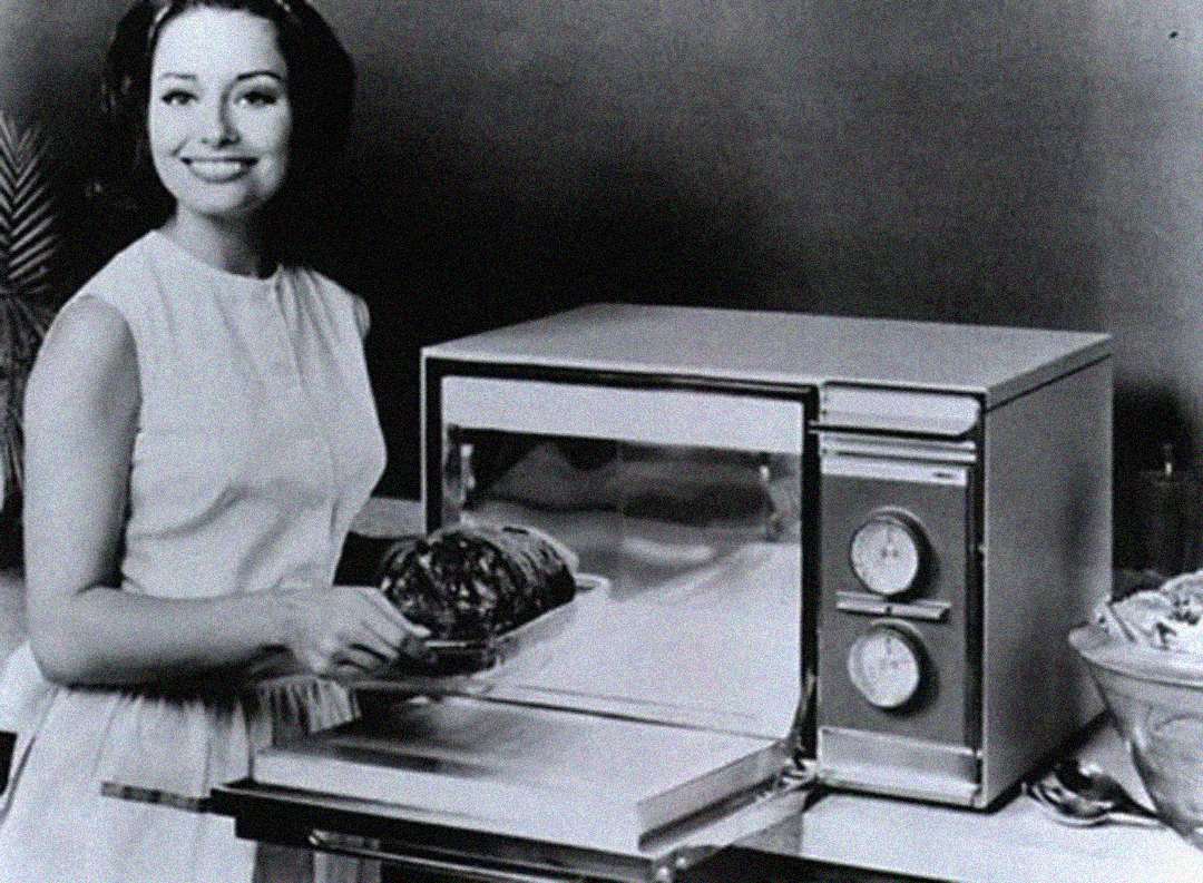 A woman in a sleeveless dress smiles while placing a roast into a vintage microwave oven, with analog dials visible on the appliance. The scene appears to be from the mid-20th century.