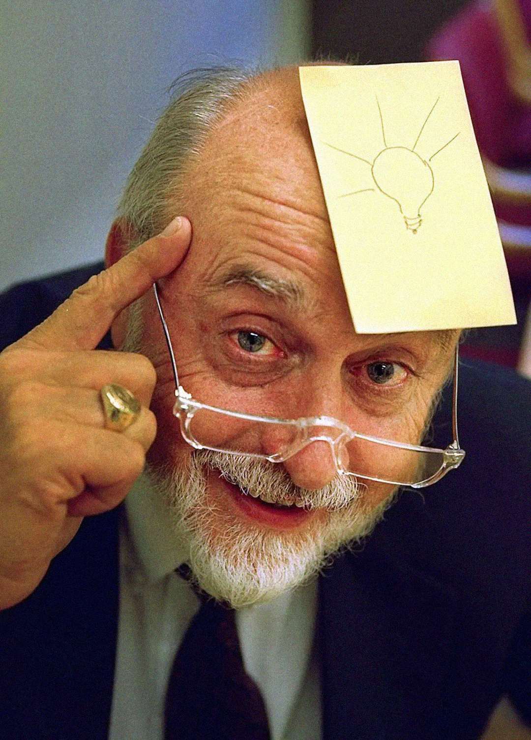 An elderly man with a beard and glasses smiles, pointing to his head where a sticky note with a light bulb drawing is placed, symbolizing an idea or inspiration.
