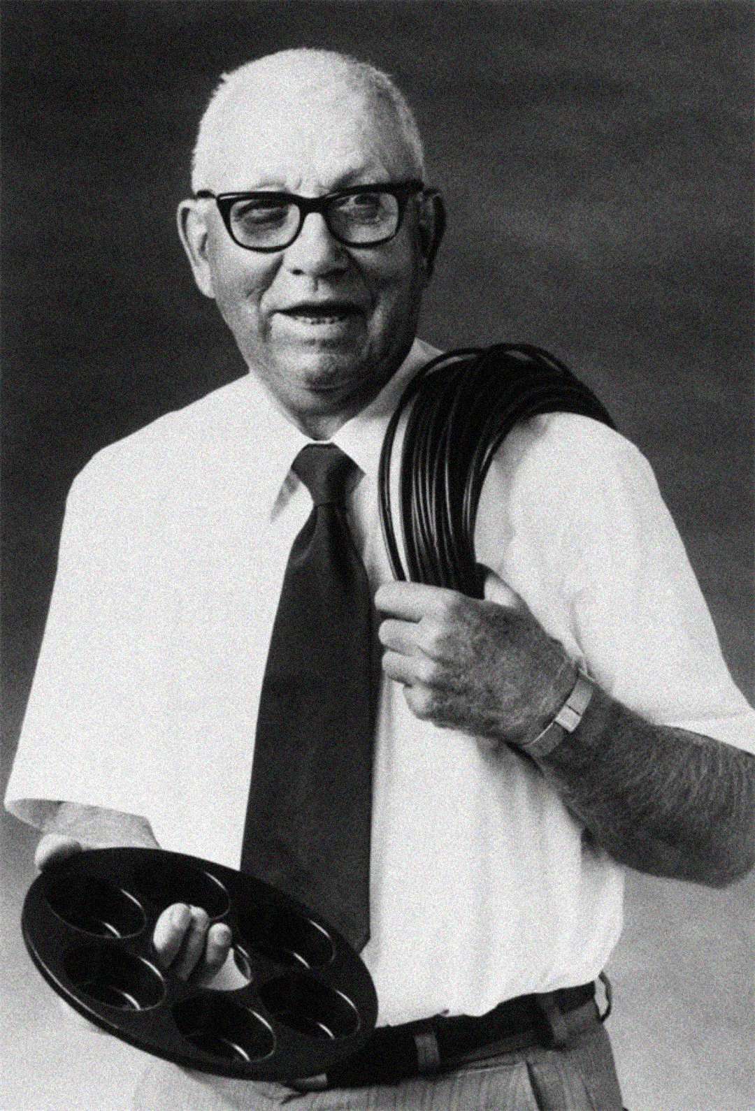 An older man wearing glasses, a white shirt, and a tie holds a circular palette in one hand and a coiled hose or cable over his shoulder, smiling at the camera in a black-and-white photo.