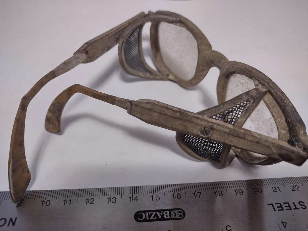A pair of old, dusty safety goggles with mesh side shields lies on a white surface next to a ruler showing measurements in centimeters and inches.