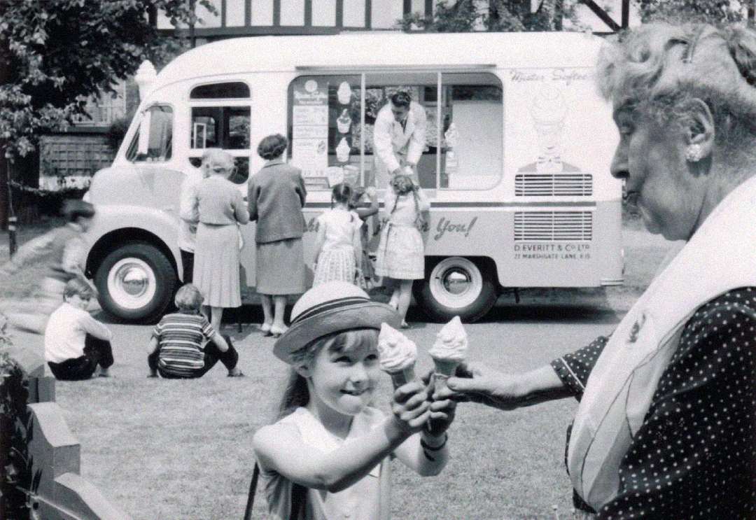 A young girl smiles as she receives an ice cream cone from an elderly woman in front of an ice cream truck, with people and children gathered around in a park-like setting.