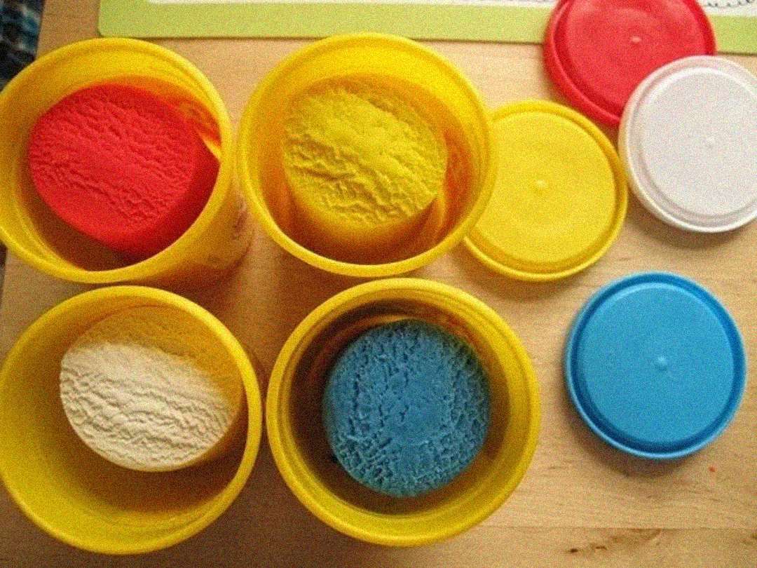 Four open yellow containers of colorful modeling clay—red, yellow, white, and blue—are shown from above on a wooden surface, with their matching lids placed nearby.