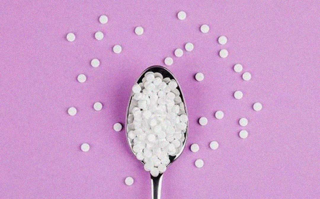 A metal spoon filled with small white round tablets is placed on a purple surface, with more tablets scattered around it.