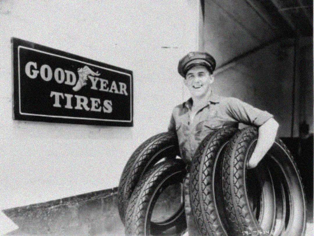 A smiling man in a uniform and cap stands holding three large tires next to a wall with a sign that reads "Goodyear Tires" in bold letters.