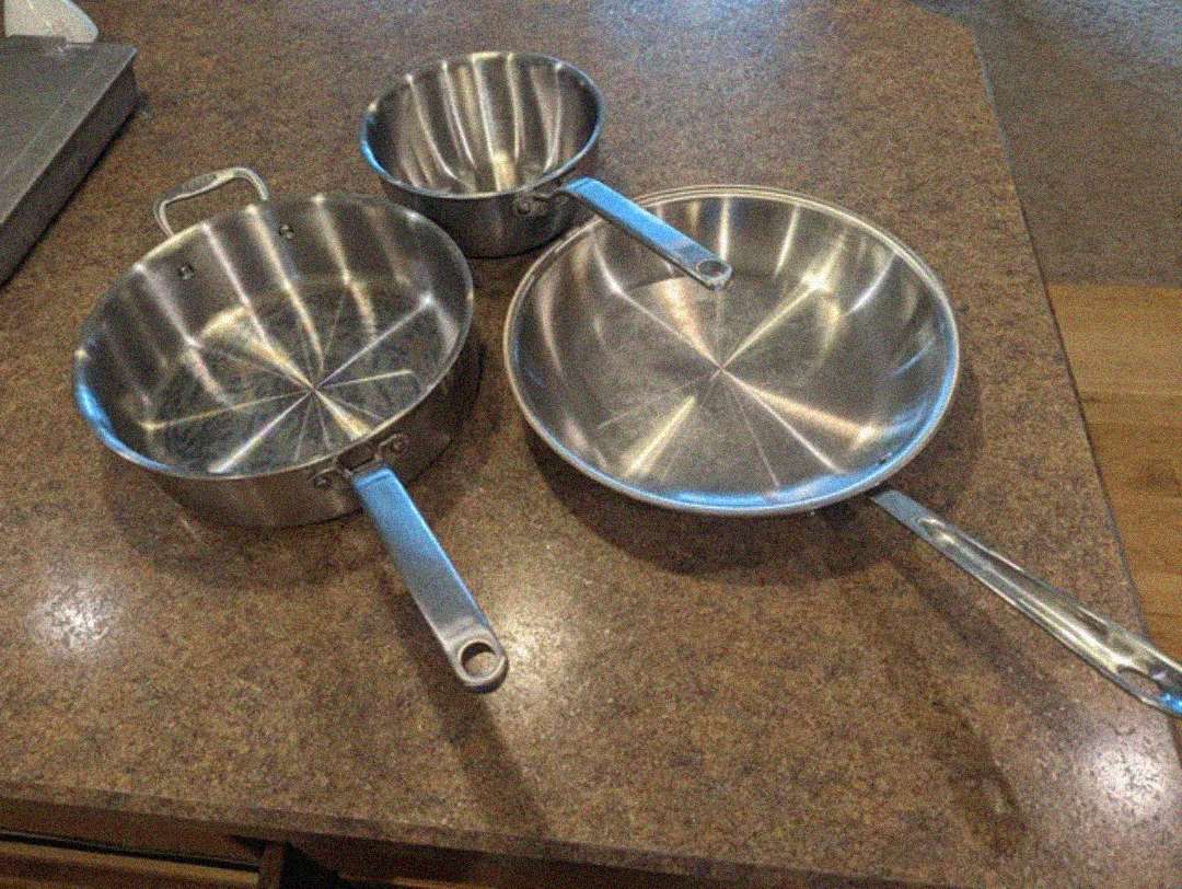 Three stainless steel pans, including two saucepans and one frying pan, are placed on a brown kitchen countertop. The pans have shiny interiors and metal handles.