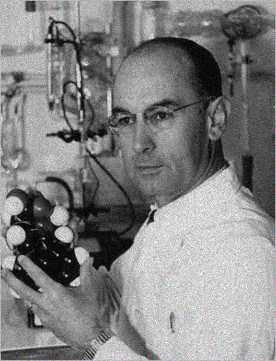 A man in a lab coat and glasses holds a molecular model in his hand, with laboratory equipment visible in the background. The image is black and white.