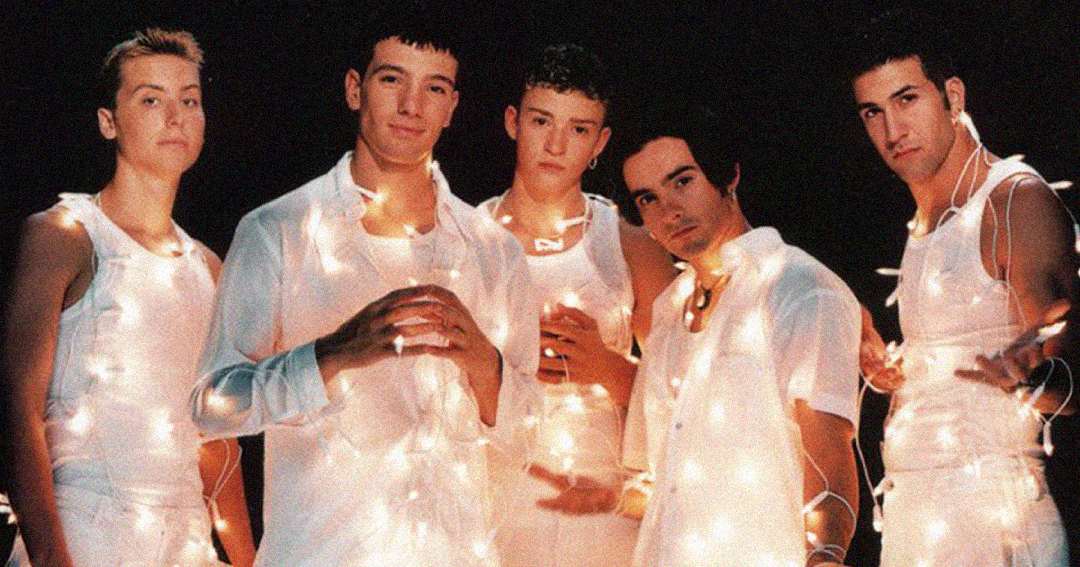 Five young men wearing white outfits are standing together against a dark background, wrapped in glowing string lights and looking toward the camera.
