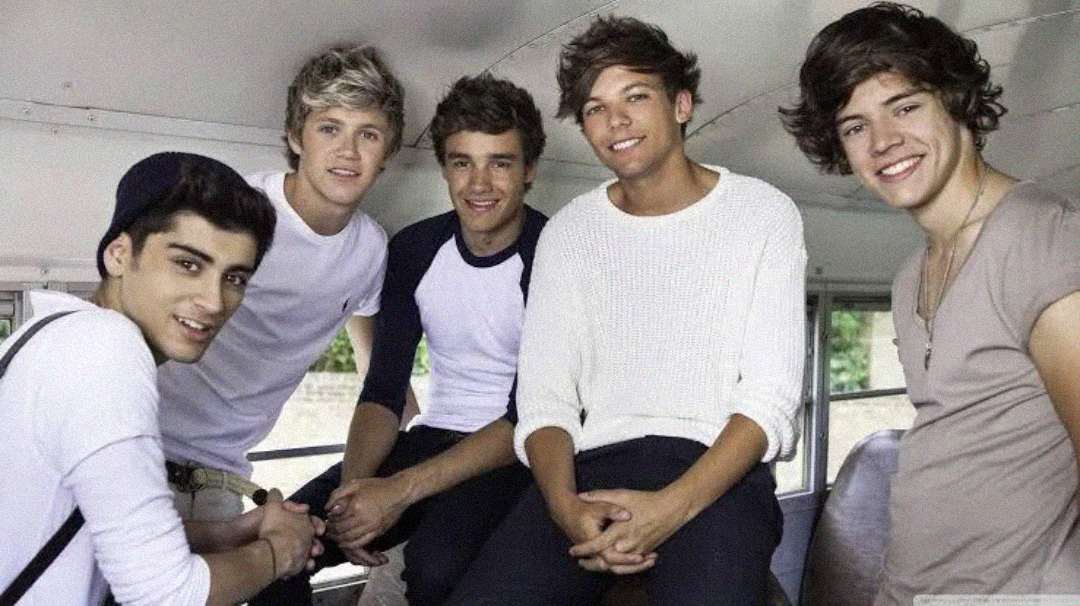 Five young men sit and smile inside a bus, casually dressed in t-shirts and jeans, posing together for a group photo with natural light coming through the windows.