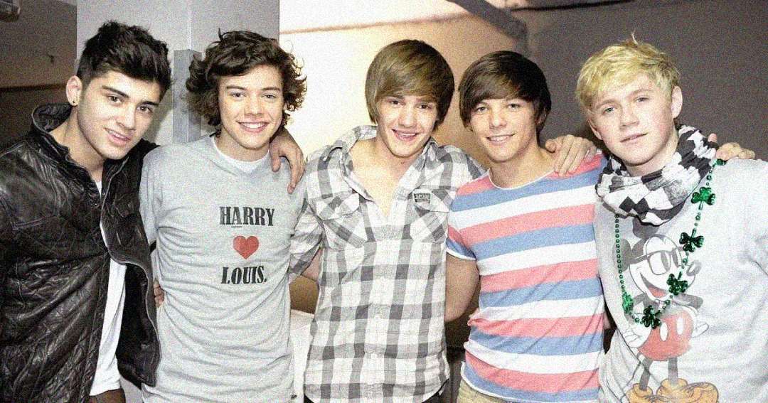 Five young men stand close together, smiling for the camera. One wears a “Harry ♥ Louis” shirt, another wears a striped shirt, and another wears a Mickey Mouse shirt with green beads. They appear happy and relaxed indoors.