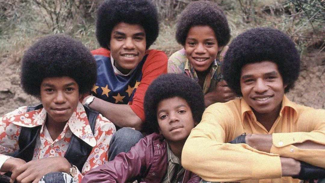 Five boys with afro hairstyles sit close together outdoors, smiling at the camera. They wear brightly colored, patterned shirts and jackets, and there is greenery in the background.