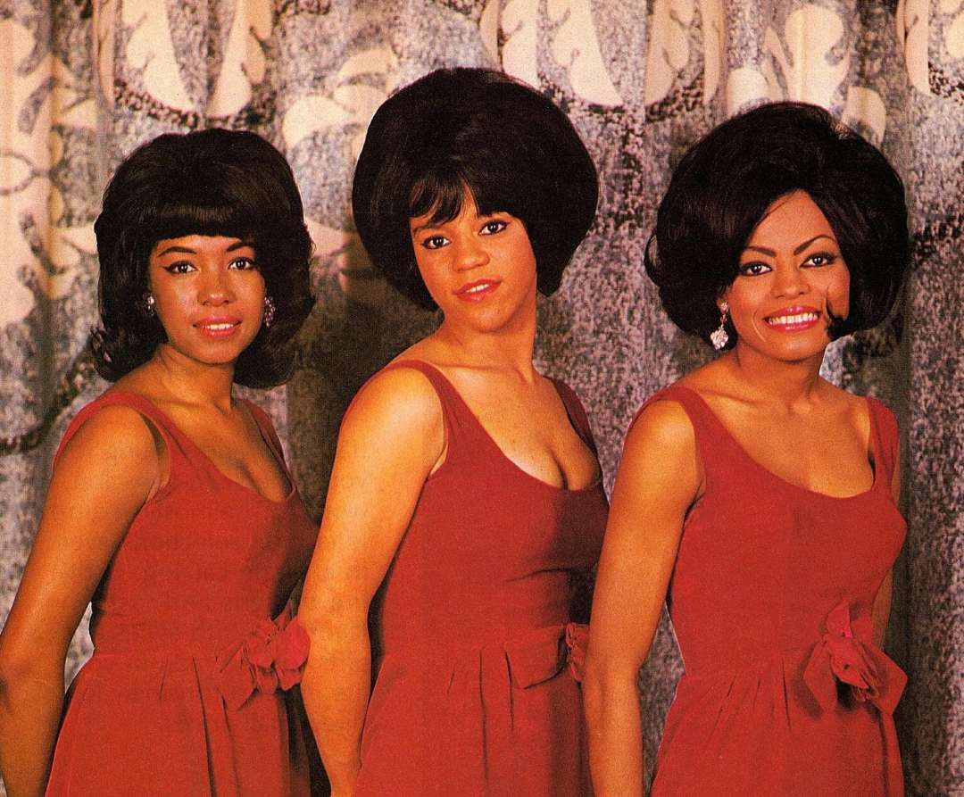 Three women wearing matching sleeveless red dresses stand side by side, smiling in front of a patterned curtain. They have styled dark hair and are posed closely together.