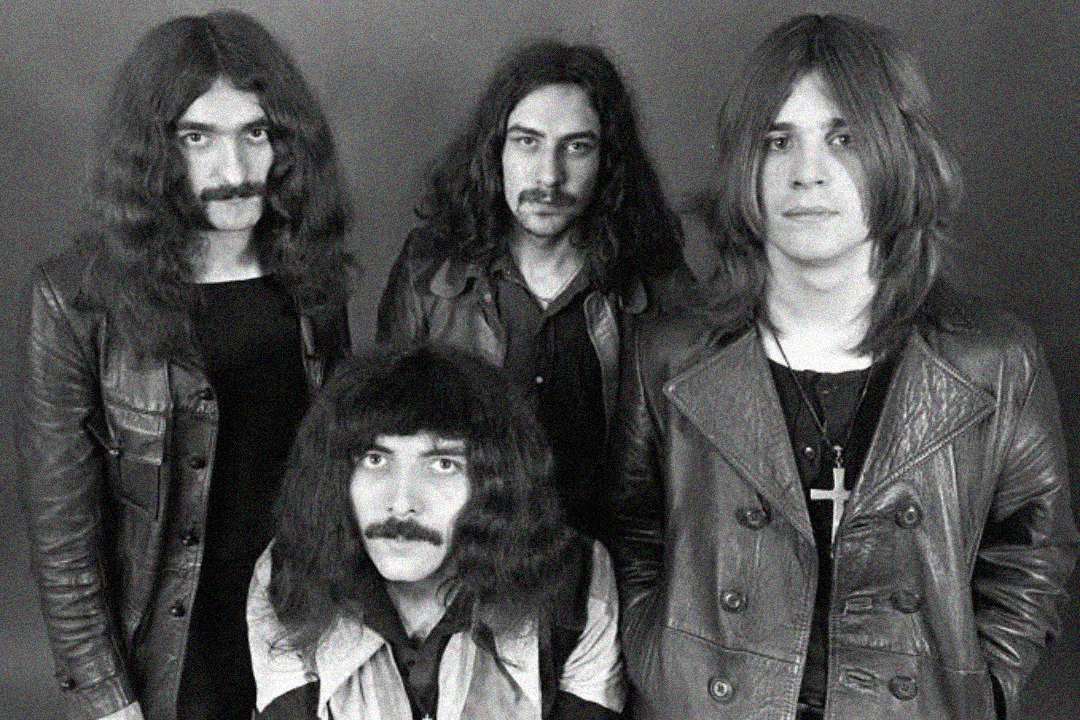 Four men with long hair and serious expressions pose together in black and white. They are wearing leather jackets and necklaces, and stand against a plain background in a vintage-style portrait.