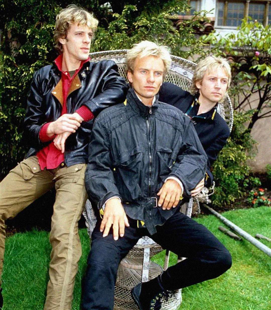 Three men with blond hair sit and stand around a wicker chair outdoors. They wear jackets and casual clothes, with green grass and trees in the background.