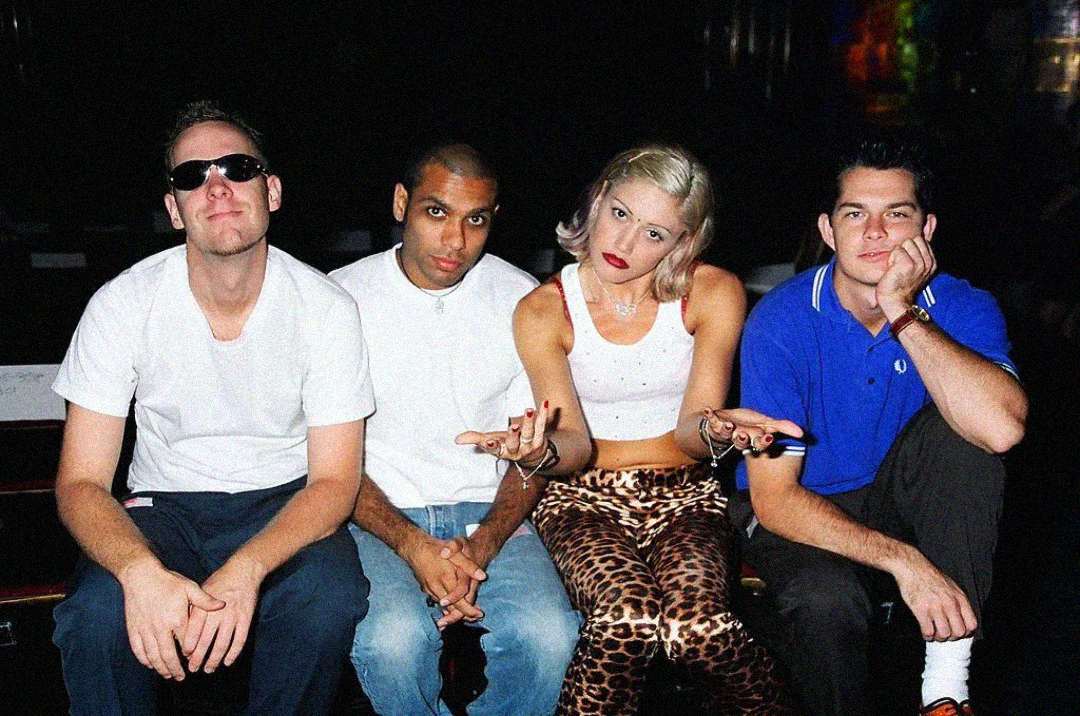 Four people sitting side by side indoors; three men and one woman. The woman wears a white top and leopard print pants, sitting between the men, gesturing with her hands. All have relaxed or playful expressions.