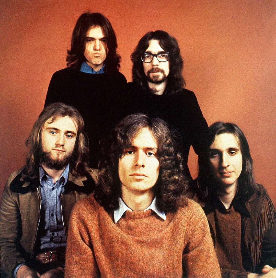 Five men with long hair and beards sit and stand in two rows against a plain reddish-brown background, all wearing casual 1970s-style clothing and looking at the camera.