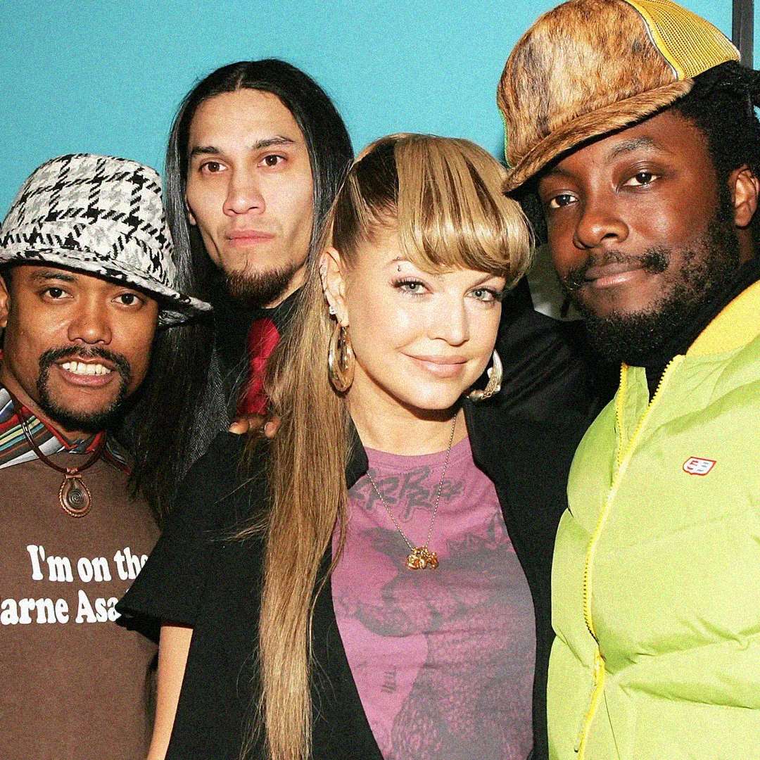 Four people stand together in a close group, smiling at the camera. The person in front has long hair and hoop earrings. The group wears colorful, eclectic clothing and hats, with a bright blue background behind them.