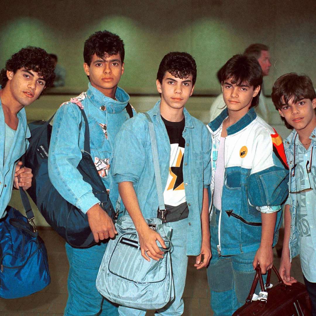 Five teenage boys wearing 1980s-style denim and casual clothes stand together, holding bags and looking at the camera in what appears to be an airport or public indoor space.