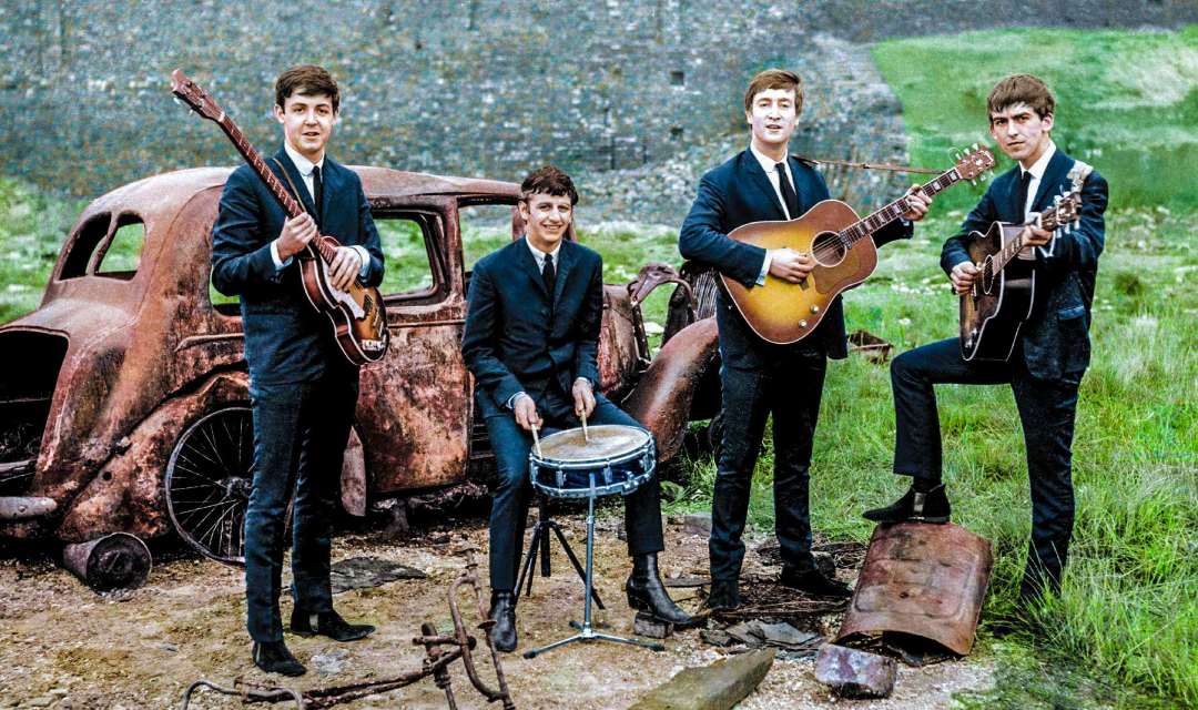 Four young men in suits play musical instruments—two guitars, a bass, and drums—standing outdoors beside a rusty, abandoned car on a grassy field with a stone wall in the background.