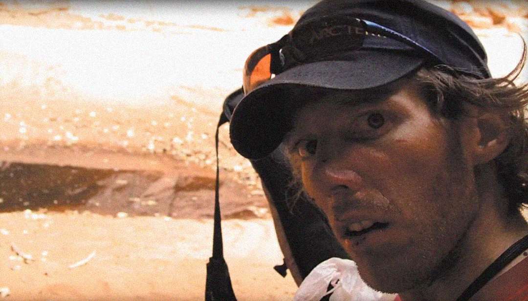 A man wearing a cap and sunglasses on his head looks at the camera with a concerned expression, standing outdoors in a sandy environment.