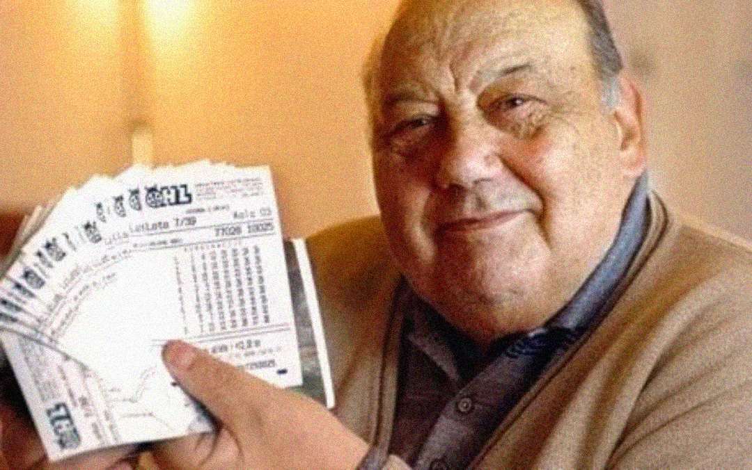 An older man smiles while holding several lottery tickets fanned out in his hand, wearing a beige sweater and sitting indoors with a warm background.