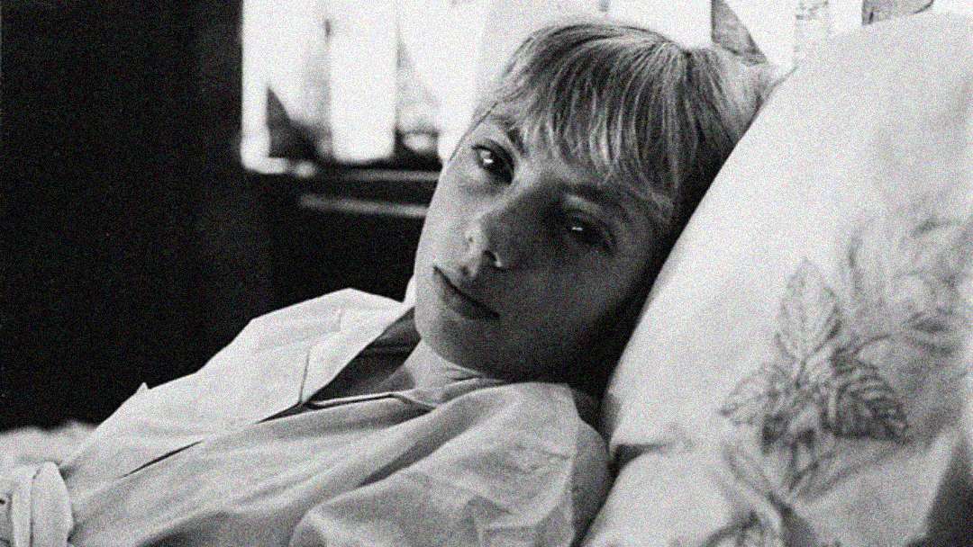 A person with short blond hair lies in bed, resting their head on a pillow with a floral pattern. They wear a light-colored shirt and look quietly at the camera. The image is in black and white.