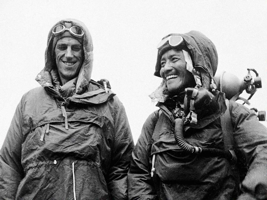 Two climbers wearing heavy jackets, gloves, and goggles stand side by side, smiling. One carries an oxygen tank on their chest. The background is bright and featureless, suggesting they are at a high altitude.