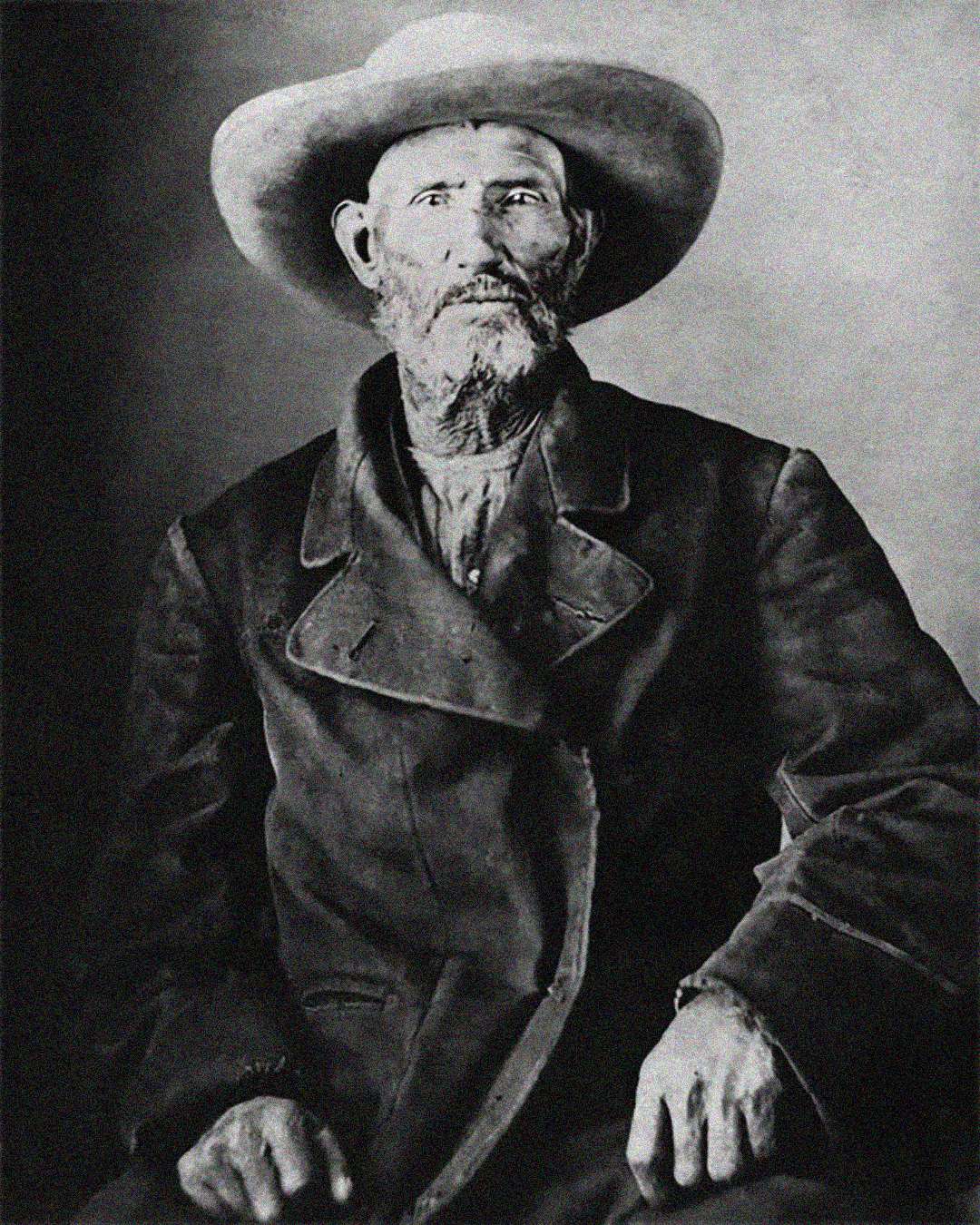 An older man with a weathered face and beard sits for a black-and-white portrait, wearing a wide-brimmed hat and a heavy, double-breasted coat, looking directly at the camera.