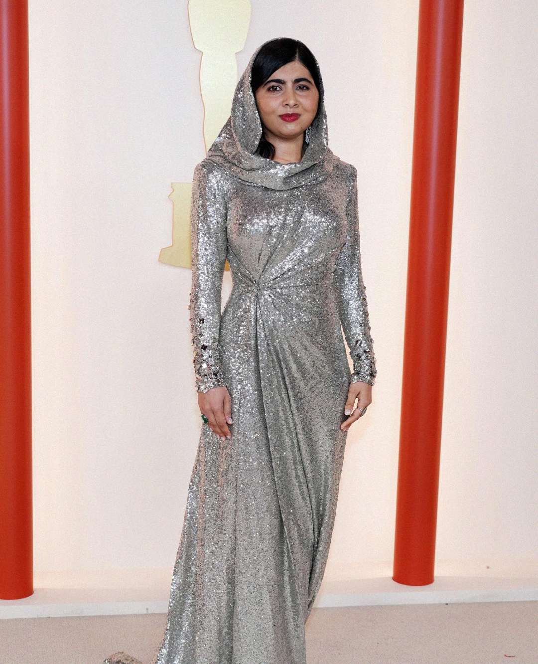 A woman stands on a light-colored floor, wearing a sparkling silver gown with long sleeves and a matching hood, posing in front of a cream backdrop with red vertical columns and a gold Oscar statuette outline.