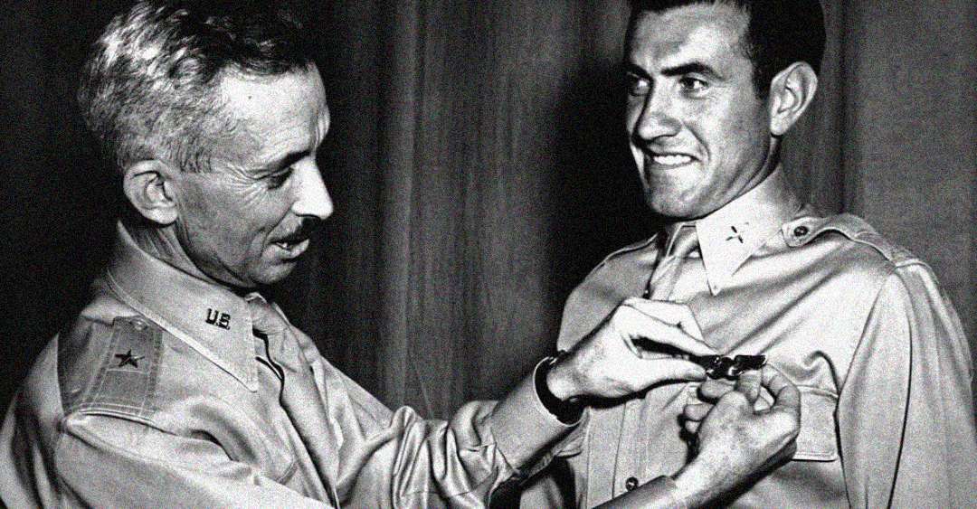 A military officer fastens a medal onto another uniformed man's chest, both wearing button-up shirts. The man receiving the medal smiles as he is honored in this black-and-white photograph.