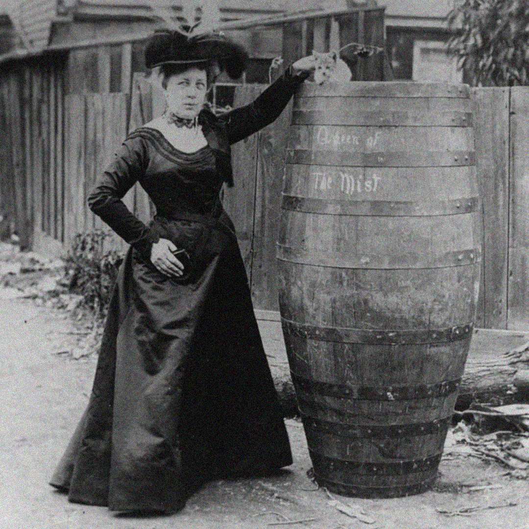 A woman in a long, dark Victorian dress and wide-brimmed hat stands outdoors, resting her arm on a large wooden barrel labeled "Queen of The Mist," near a wooden fence.