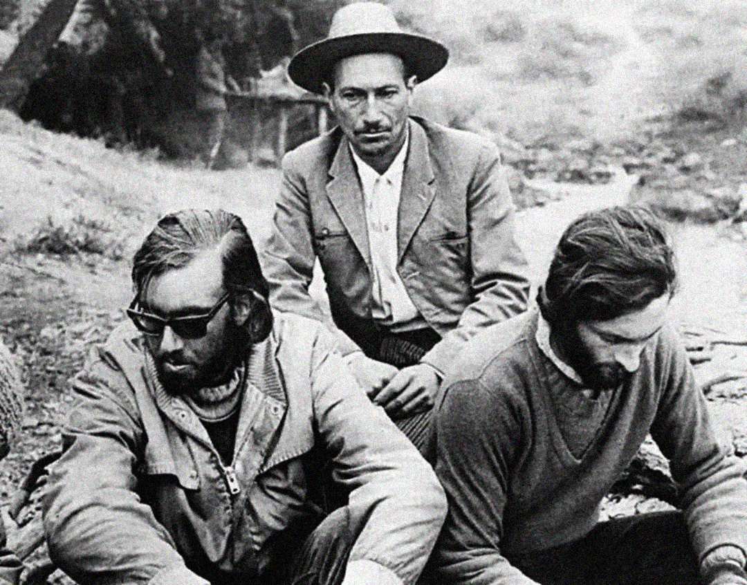 Three men sit outdoors in a black-and-white photo; one wears sunglasses, another a wide-brimmed hat, and the third looks down with long hair and a beard. The setting appears natural and informal.