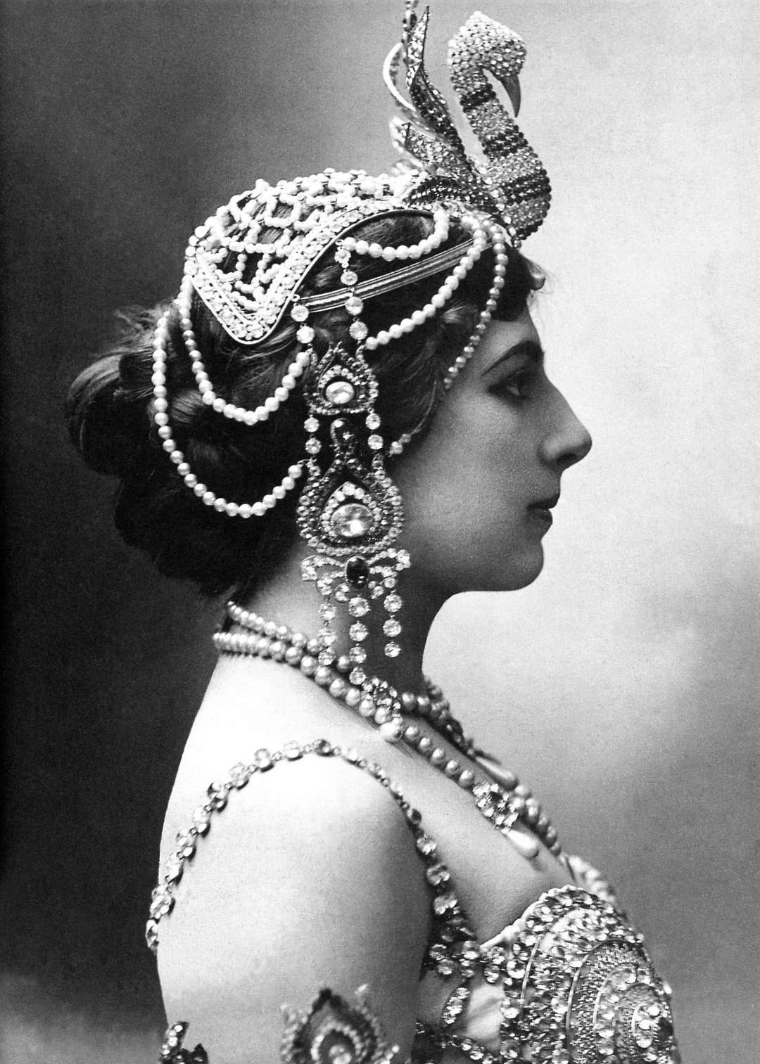 A woman in elaborate costume and jewelry, including pearl strands, a beaded headdress with a decorative bird, and ornate earrings, poses in profile against a neutral background.