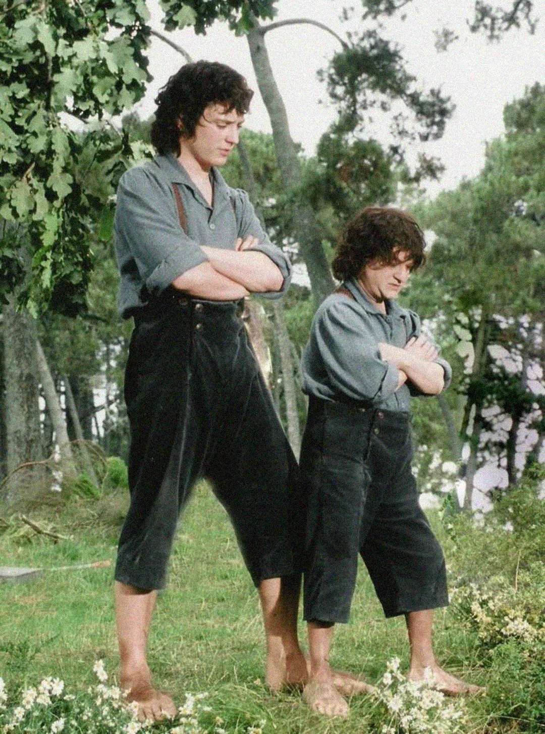 Two men with curly hair, wearing similar outfits of blue shirts and black pants, stand barefoot outdoors with arms crossed. One man is much taller than the other; they appear to be in a forested area with grass and wildflowers.