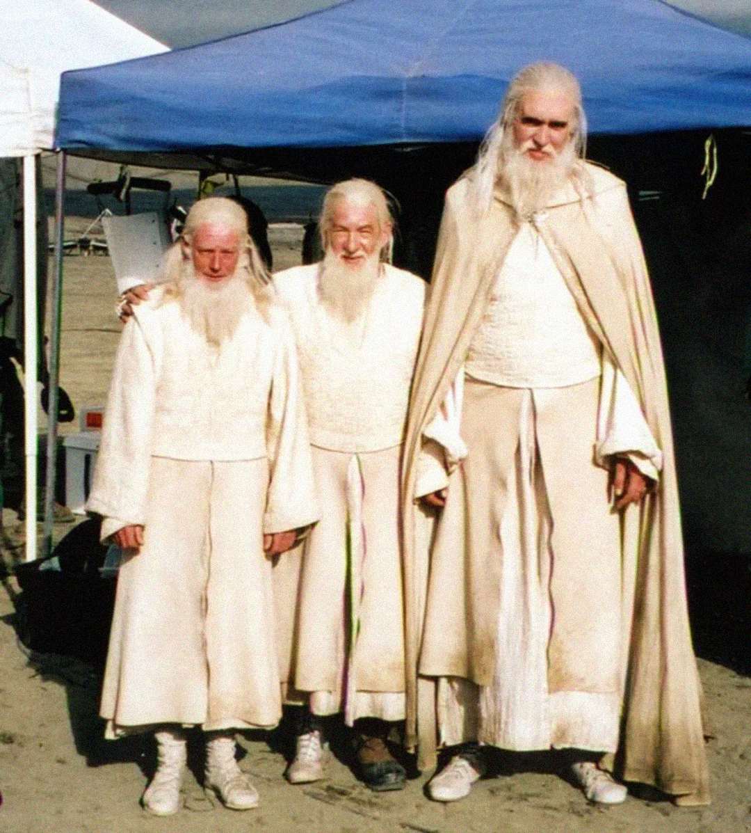 Three people dressed in long white robes and fake white beards stand under a blue canopy outdoors, posing for a photo. The tallest person is much bigger than the others and wears a long cape.