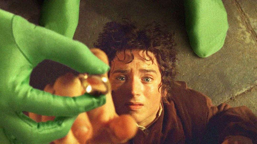 A person with curly brown hair and a distressed expression looks up as a hand in a green glove holds a gold ring near their face, with green-clad feet visible in the background.